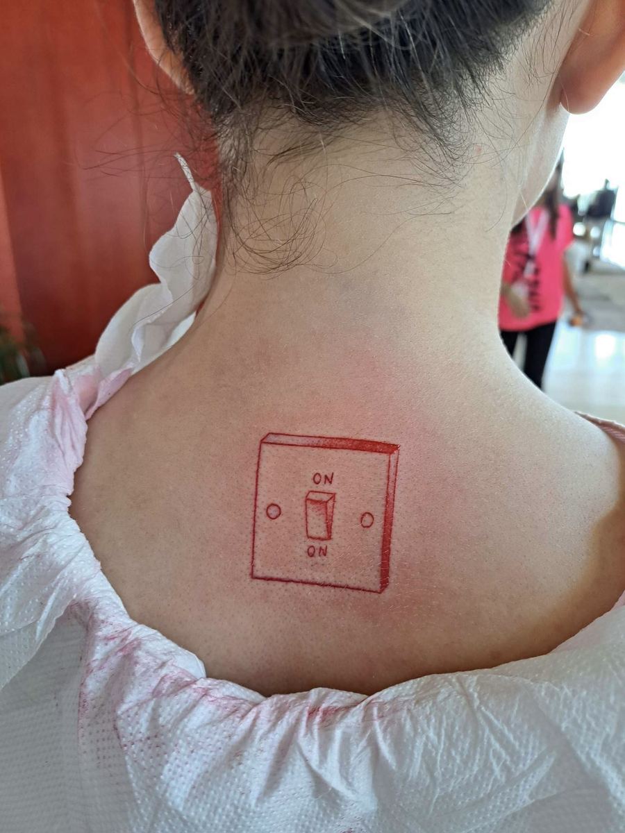 A red tattoo of a light switch on the back of Ismini's neck, labelled ON at the top and ON at the bottom